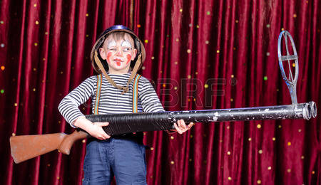 Young-boy-wearing-clown-make-up-and-military-helmet-standing-on-stage-with-red-curtain-aiming-over-s